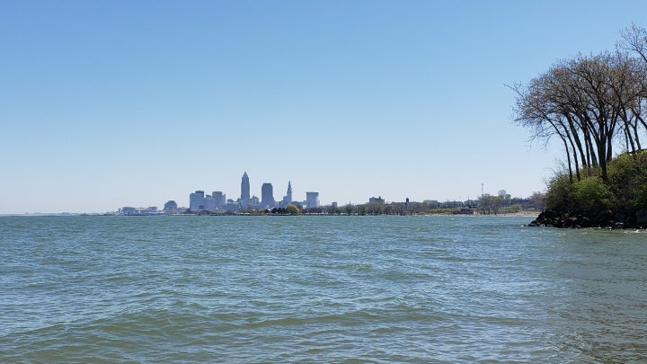 A calm lake with gentle waves is in front of the Cleveland skyline, visible in the distance. A cluster of trees stands on the right shoreline.