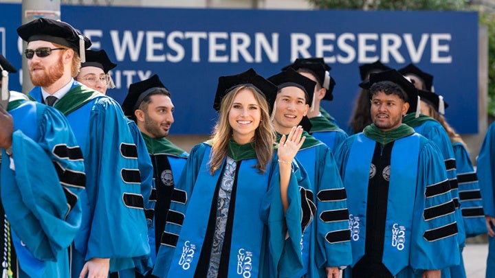 A CWRU grad waves as she walks with a procession to a commencement ceremony and passes the CWRU sign on Adelbert Road and Euclid Avenue