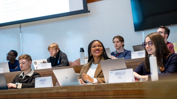 Seven students sit in a tiered classroom in the Peter B. Lewis Building. Two students smile and interact with each other while the others focus on their laptops. 