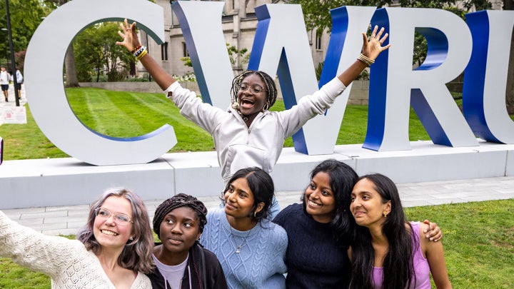Five CWRU students pose for a selfie photo as another student leaps with their arms up behind them in front of the CWRU letters on campus