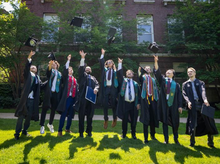 A group of undergraduate students wearing graduation attire jump in unison while throwing their caps in the air