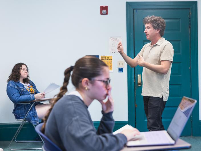 A classroom setting with a teacher standing and speaking. Two students, one focused on papers and the other on a laptop, listen attentively.