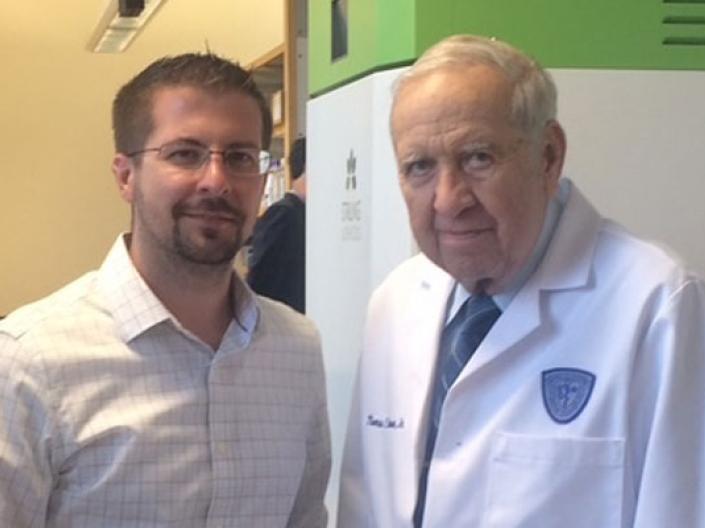 Two men standing side by side, one on left wearing dress shirt and on right white lab coat