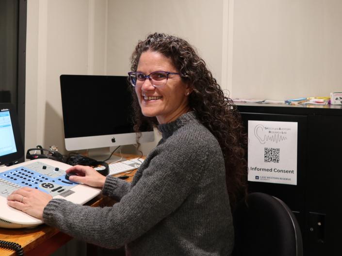 A woman sits with hearing assessment equipment in a sound-isolated booth