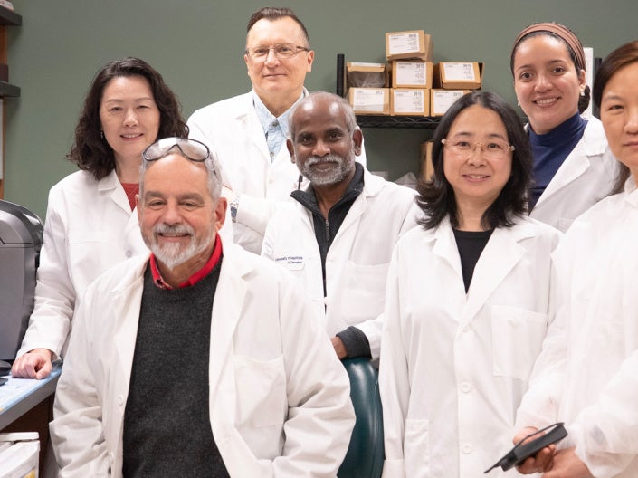 Seven researchers in white lab coats pose together inside a biomedical laboratory. Shelves lined with binders and lab supplies surround the group, while a computer monitor on the left displays colorful microscopic cell images. 