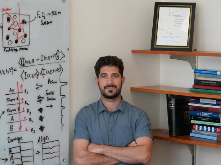 A researcher stands beside a whiteboard covered with diagrams, equations, and notes about imaging, scattering, and polymers. On his right, there are shelves filled with scientific textbooks and framed research awards.