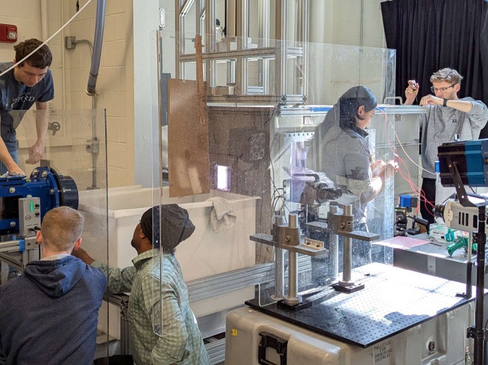 Researchers prepare a hypervelocity water-entry experiment, with the HOPLITE two-stage light gas gun at left, a water tank at right, and high-speed imaging cameras aimed through the tank’s clear glass wall to capture the projectile’s interaction with water.