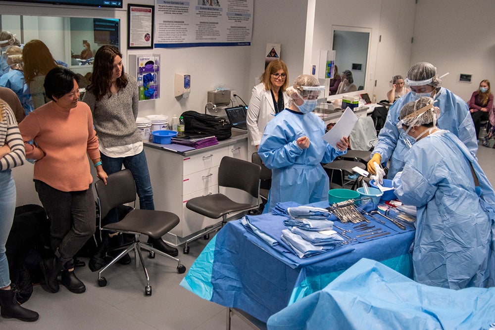 Students on left watch as nursing students in full scrubs perform a mock lab surgery