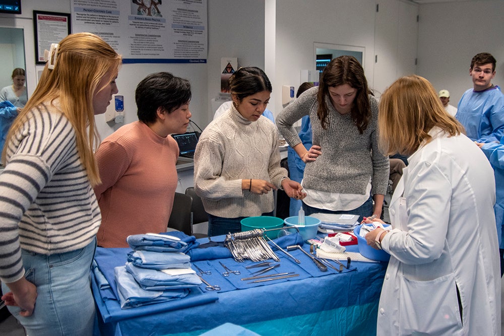 4 Female students look over a table filled with nursing instruments while a white coat instructor oversees