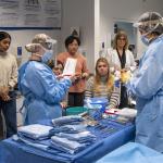Nursing students in blue scrubs count surgery items in a mock simulation while other students watch