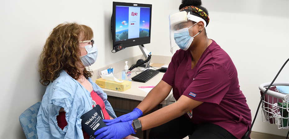 Primary care nurse taking a patient's blood pressure. Both are wearing face masks.