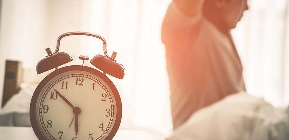 Alarm clock in foreground and a man waking up and stretching in bed in the background.