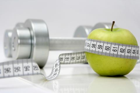 Stock photo of a measuring tape wrapped around a green apple with a set of free weights in the background.
