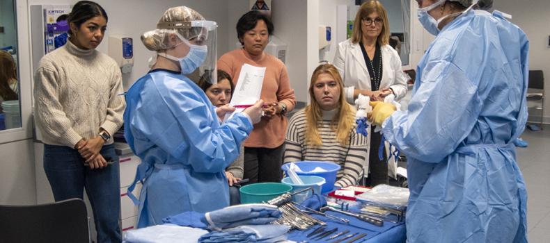 Nursing students in blue scrubs count surgery items in a mock simulation while other students watch