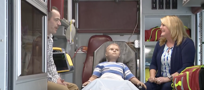 Tom Baum, left, sits in the ambulance simulator with a mannequin, center, and reporter Monica Robbins, right.