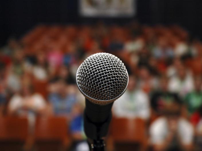 Stock image of a closeup on a microphone with an audience in the background.