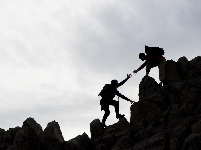 Silhouette image of two people climbing rocks or a mountain and one person is reaching down to help the other.
