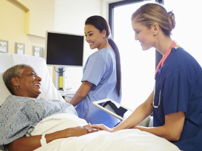 Stock photo of a older African American female patient in a bed talking to two female nurses who are checking vital signs.