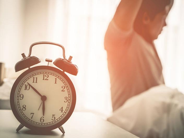 Alarm clock in foreground and a man waking up and stretching in bed in the background.