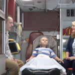 Tom Baum, left, sits in the ambulance simulator with a mannequin, center, and reporter Monica Robbins, right.