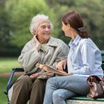 An older woman sits on a park bench outside with a younger woman sitting next to her on the right
