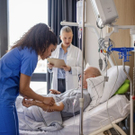 Stock image of a nurse in blue scrubs and nurse in white coat helping a patient in bed