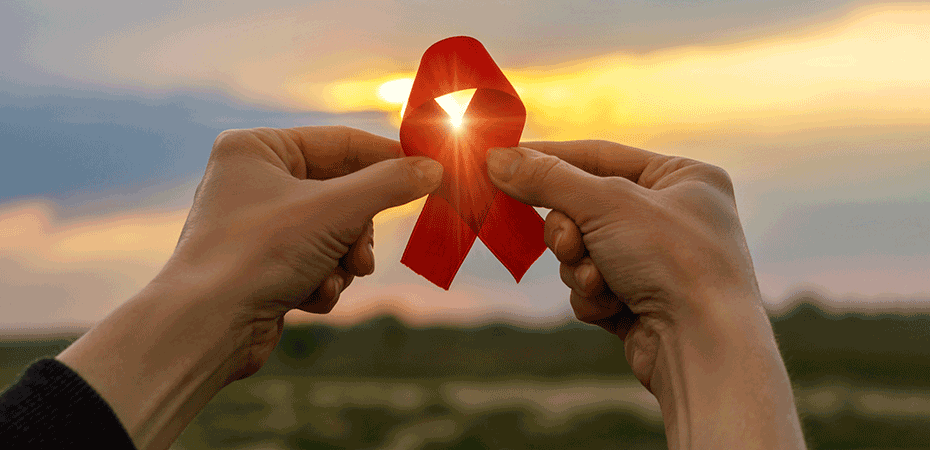Hands holding red HIV awareness ribbon in front of a sunset.