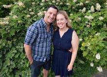 A man in plaid shirt at left with a woman in blue dress at right with green foliage behind them