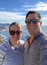 A man and woman both wearing sunglasses smiling with the coastal water behind them