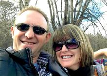 A man and woman, both in sunglasses, smiling with trees behind them