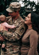 A man in military uniform holding a baby while a woman stands beside them smiling