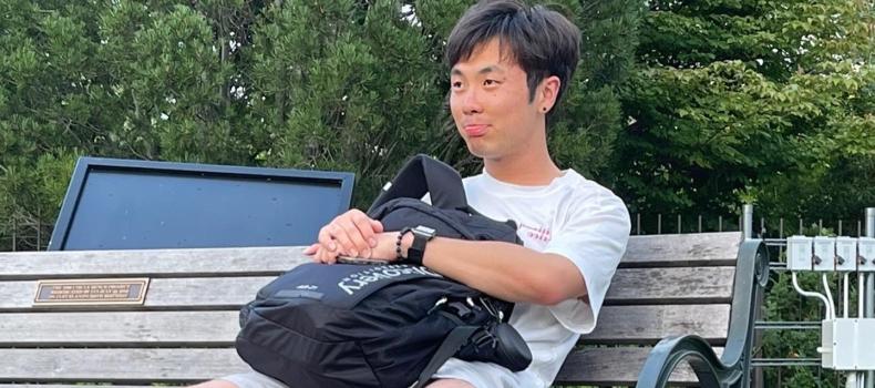 BSN student David Kim Koh sits on a bench holding his backback while smiling