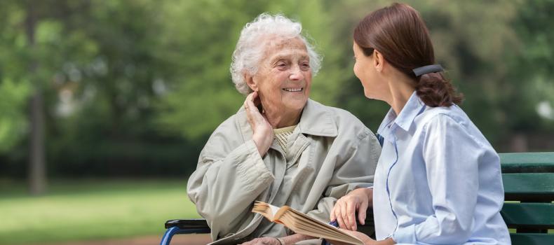 An older woman sits on a park bench outside with a younger woman sitting next to her on the right