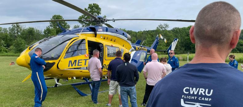 A group of participants at CWRU Flight Camp 2024 gathered around a yellow and blue MetroHealth medical helicopter in a grassy field.