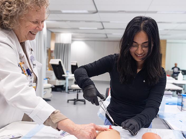 A smiling nursing student in black gloves practices a procedure, likely suturing, on a medical training manikin while a smiling female instructor in a white coat guides her technique.