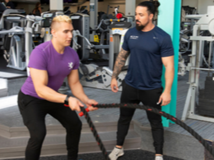 Male personal trainer with male client in purple shirt using battle rope
