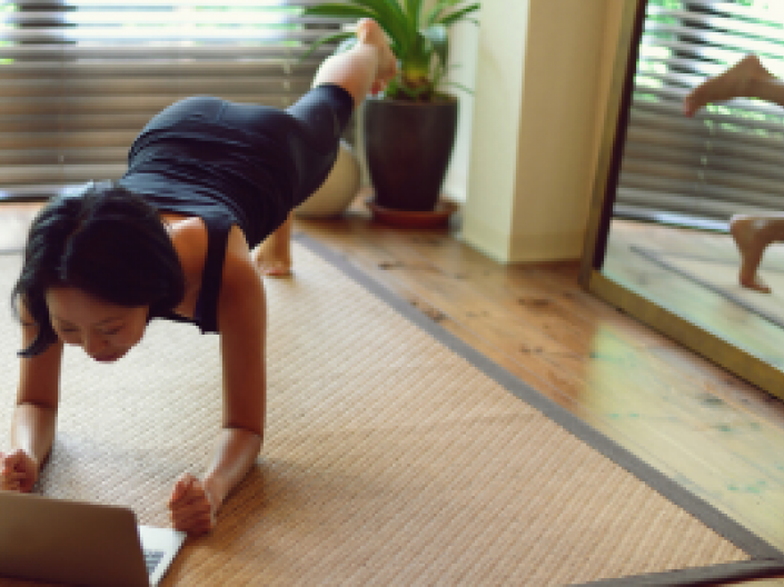 Woman in plank pose on floor of bedroom looking at computer