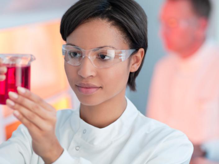 A woman examining a beaker wearing a lab coat and goggles