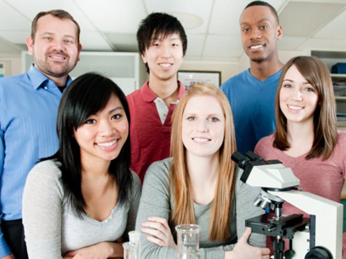 A group shot of 6 people in a lab standing behind a microscope