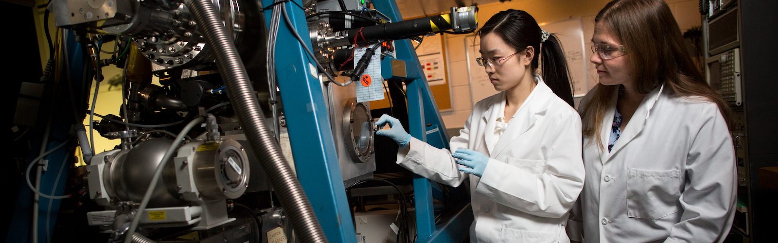 Two students in lab coats closely examine a machine 