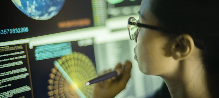 Photo of a young Asian woman, pointing at, and learning from, global data displayed on a large computer monitor.