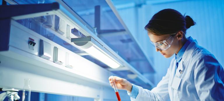 A female presenting scientist in lab coat and PPE looks at a test tube in a lab setting