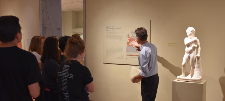 A group of students observe a professor speak and point at a map beside a classical art statue