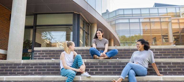 Three femme students of differing ethnicities sit and chat on the steps of the Mandel Center