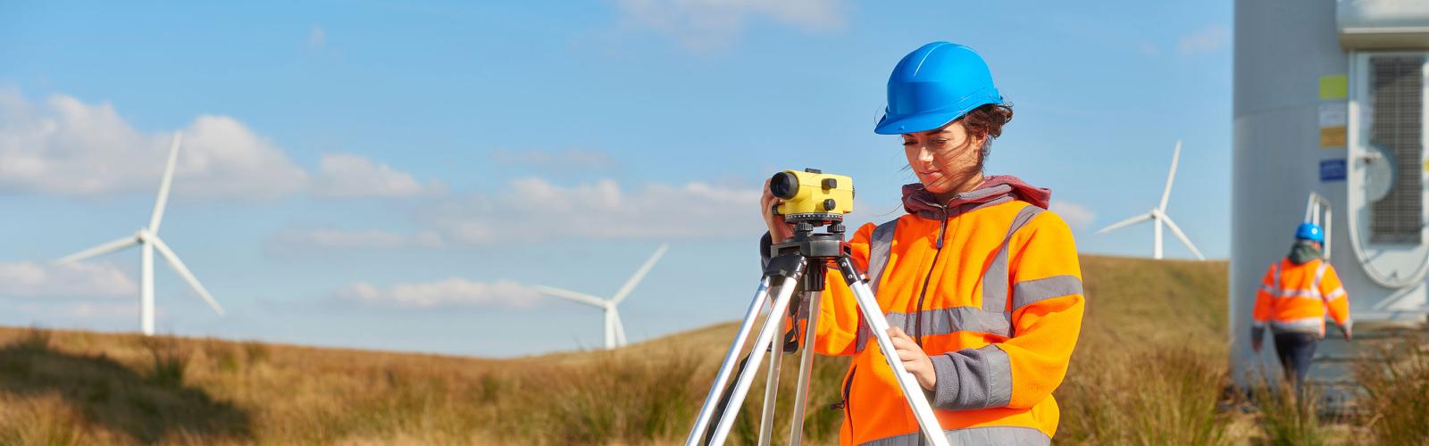 Two wind farm engineers working around the base of a wind turbine , they are wearing orange hi vis jackets and blue hard hats . one is male , one is female. In the background wind turbines can be seen across the landscape. In the foreground a female engineer uses a builder's level whilst her colleague accesses the entrance to the turbine.