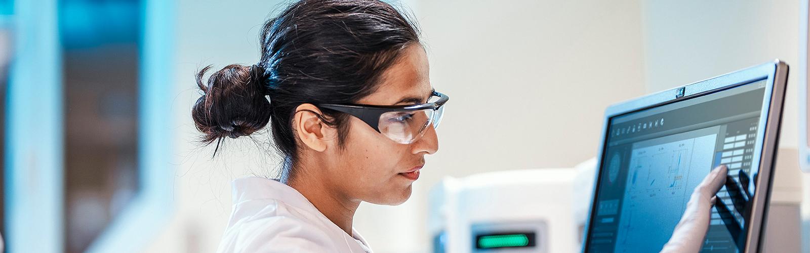 A researcher wearing safety glasses looks intently at data displayed on a computer screen in a lab.