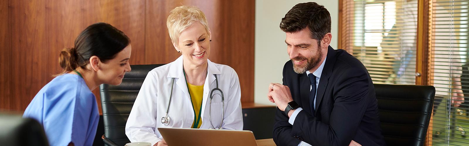 Three professionals—a nurse, a doctor, and a businessman—sit together looking at a laptop in an office.