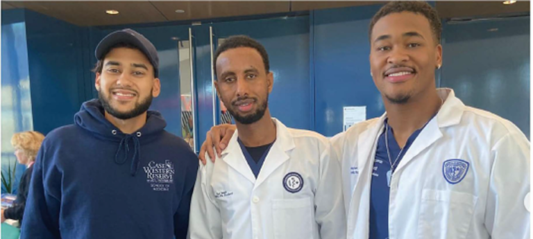 Three masculine students of color pose wearing CWRU branded doctor coats and scrubs