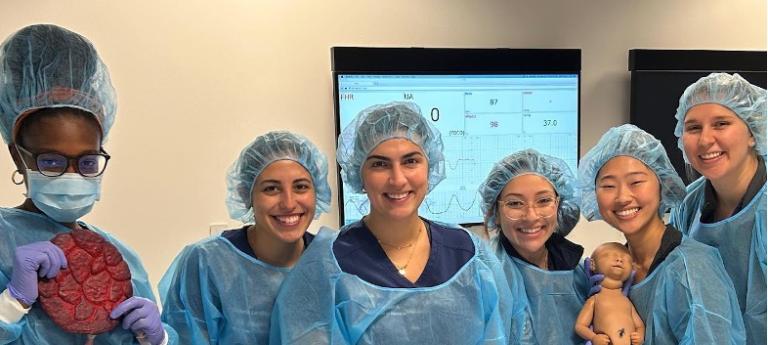 Six femme students of various ethnicities pose in surgical gear with a practice baby and patient