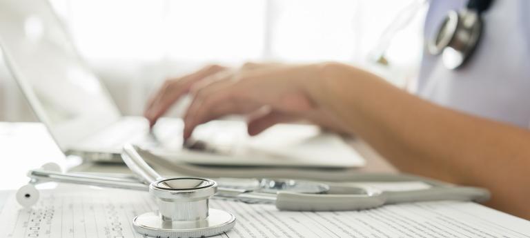 A medical professional's hands typing on a laptop, with a stethoscope resting beside it.